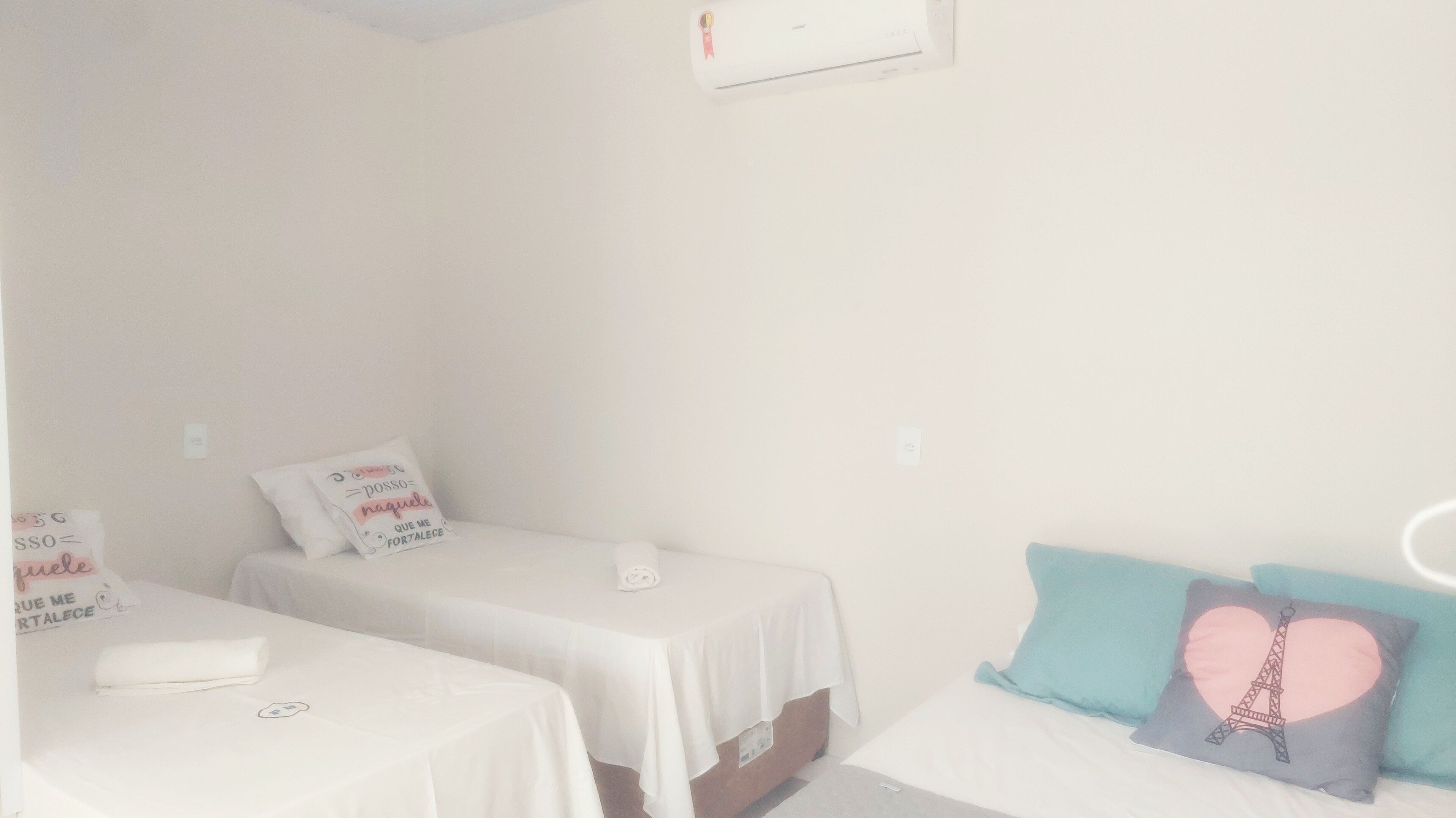 A simple, clean room with two single beds, each with a white bedspread and a pillow with text. There is a small table with a white tablecloth and a folded white towel. The room has white walls and a small window with a curtain.