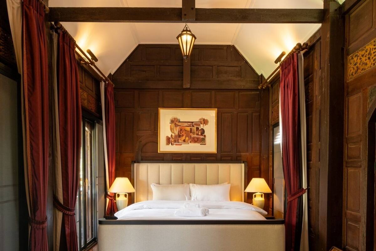 A large bed with a headboard, two bedside lamps, and a hanging light fixture in a room with wooden walls and a sloped ceiling.