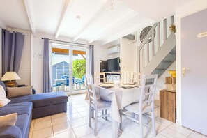 A modern living room with a sofa, dining table, and chairs, a kitchen area with a TV, and a staircase leading to another level.