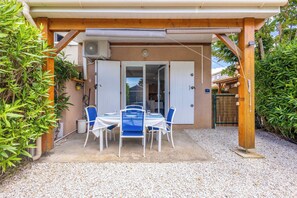 Une terrasse extérieure avec une table et des chaises, un espace couvert doté d’une structure en poutres de bois, et une vue sur l’intérieur de la maison.