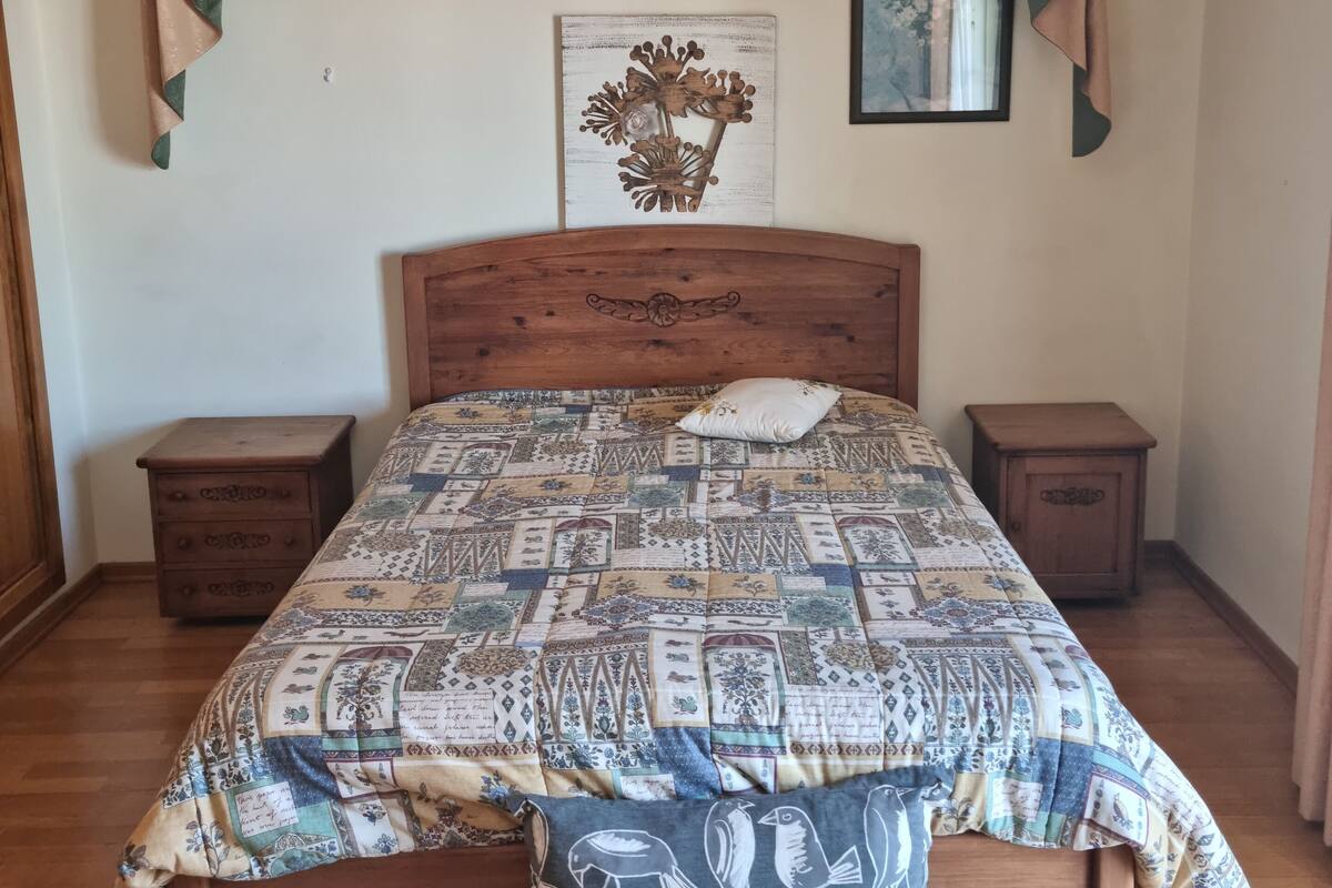 Ein Schlafzimmer mit einem hölzernen Bett, zwei Nachttischen, einer Pendelleuchte, einer Wanddekoration und einem Fenster mit Vorhängen.