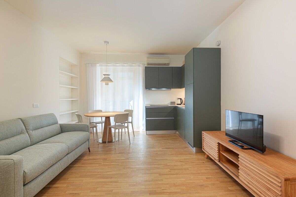 Una sala de estar moderna con un sofá, una mesa de comedor con sillas, un televisor sobre un mueble de madera y una cocina con armarios y aire acondicionado.