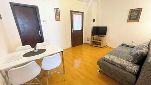 Un salon comprenant un canapé, un téléviseur posé sur un meuble, une table à manger avec des chaises et une porte avec un rideau.