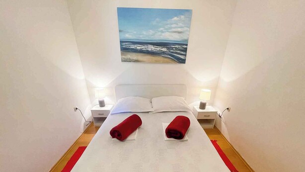 Une chambre d’hôtel avec un lit, deux tables de chevet, une petite table, un tableau représentant une scène de plage et un tapis rouge sur le sol.