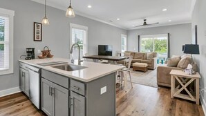 A modern kitchen with a central island, a dining area with a sofa, and a living room with a TV. - The Beacon, Steps From The Beach (Carolina Beach)