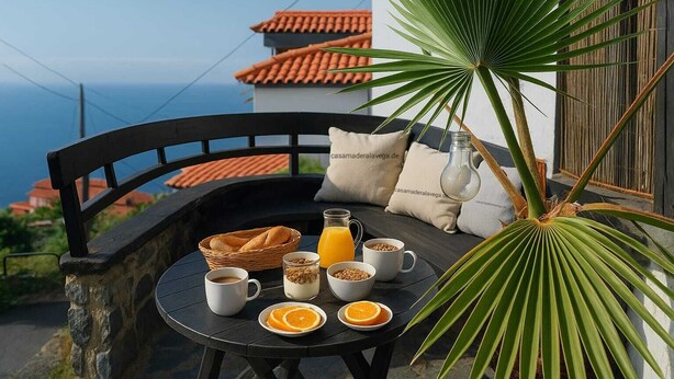 A table set with breakfast items including bread, coffee, and orange slices, with a view of the sea and a building with red tiles in the background. - Casa Madera 2 (La Vega)