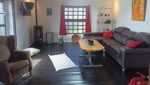 A cozy living room with a dark wooden floor, a brown armchair, a grey sofa with red cushions, a small wooden table with a bowl, and a window with red curtains. - Casa Madera 2 (La Vega)