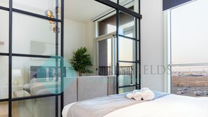 Habitación de hotel con un ventanal grande, una cama con cabecero gris, una planta en maceta y vistas a un estacionamiento.