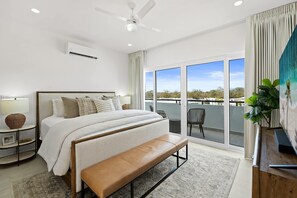 Une chambre moderne avec un grand lit, un banc, un ventilateur de plafond et une vue sur l’extérieur grâce à de grandes fenêtres.