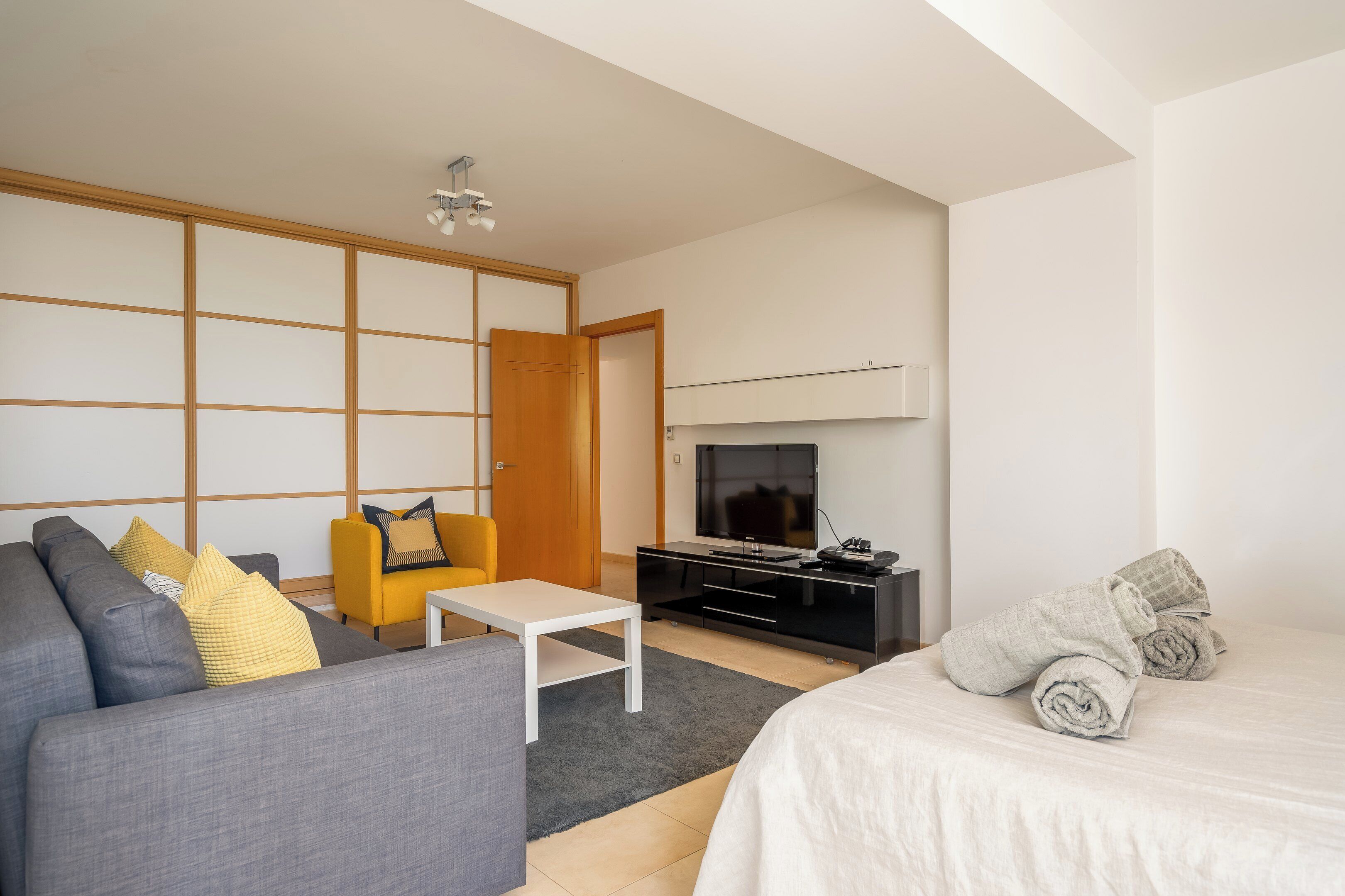 Ein modernes Hotelzimmer mit grauem Sofa, einem kleinen weißen Tisch, einem gelben Sessel, einer schwarzen Unterhaltungsanlage, einem Bett mit weißer Bettwäsche und einem großen Fernseher.
