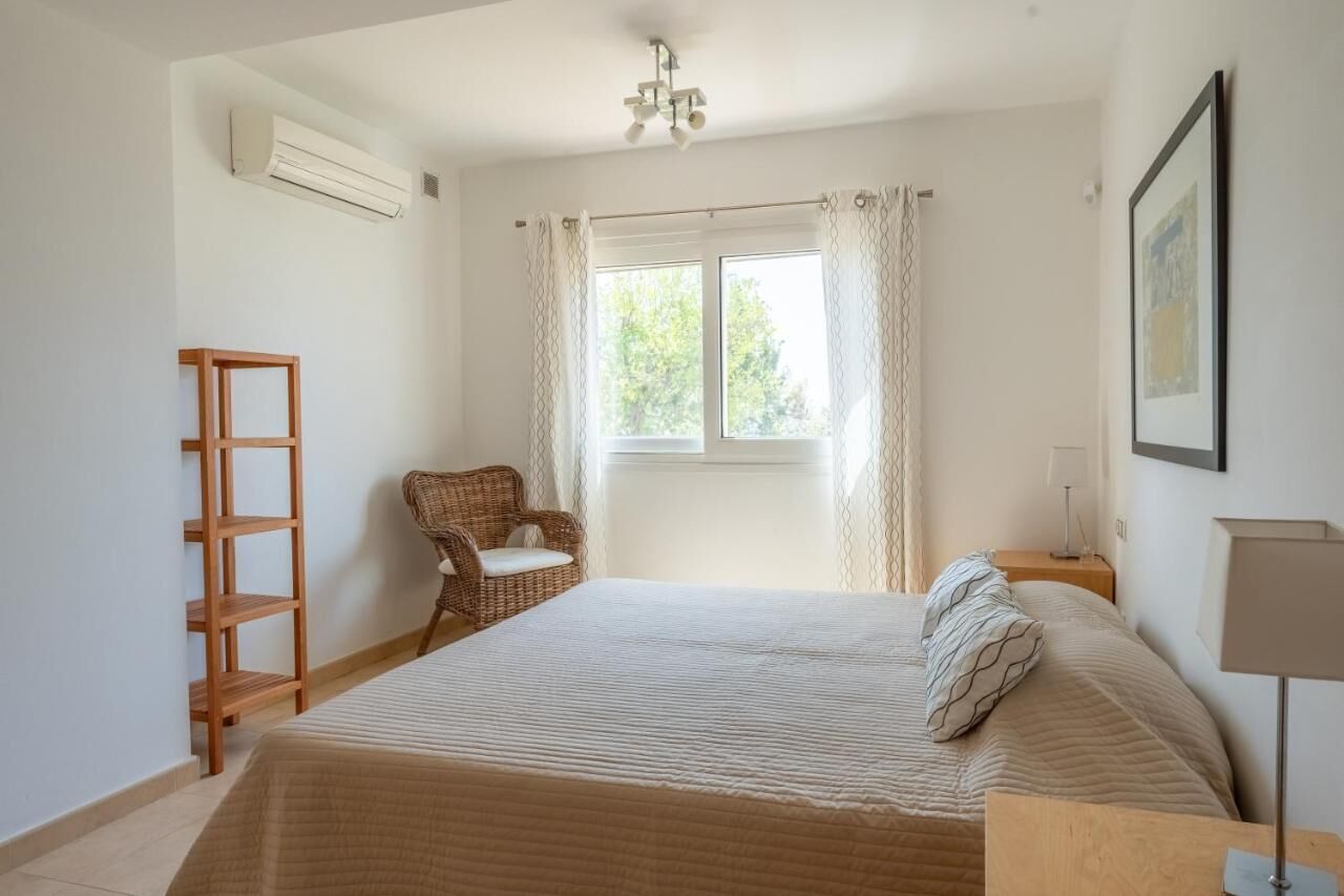A bedroom with a bed, a chair, a wooden shelf, a window with curtains, and a wall-mounted air conditioner.