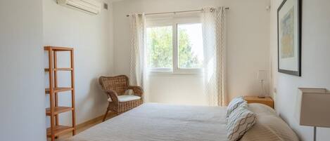 A bedroom with a bed, a chair, a wooden shelf, a window with curtains, and a wall-mounted air conditioner.