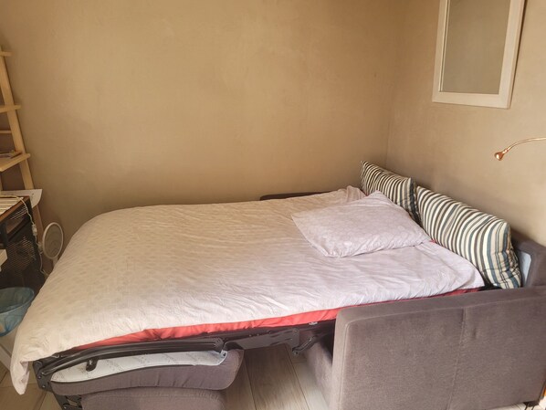 Una cama compacta con colchón blanco y un detalle en rojo, cabecera tapizada gris y almohada a rayas.