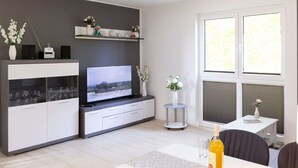 A modern living room with a flat-screen TV, white cabinetry, a small round table with a vase, and a large window.