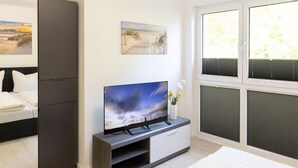 A modern hotel room with a bed, a TV on a stand, a painting on the wall, and a window with blinds.