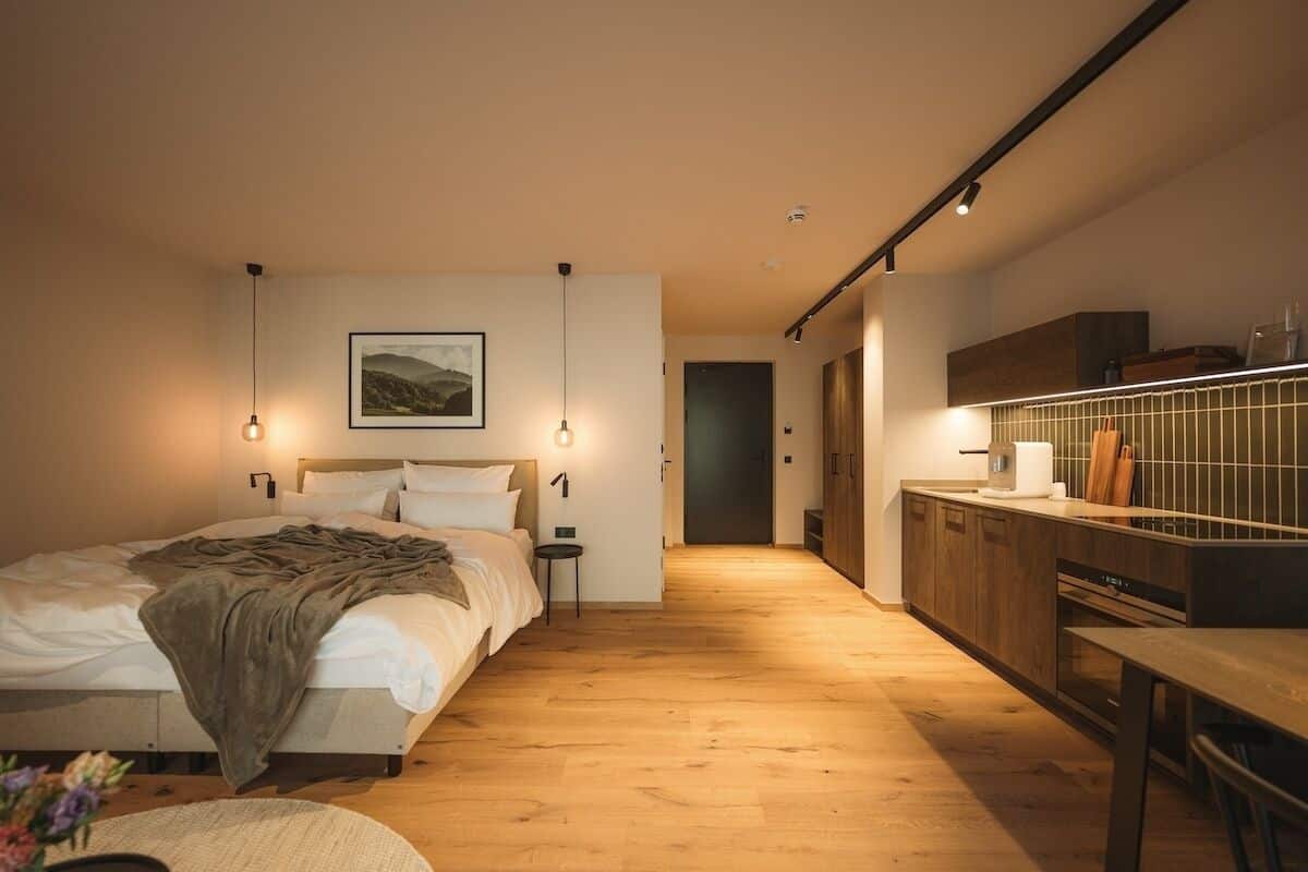 Un dormitorio moderno con piso de madera, una cama con una colcha, lámparas de noche y una mesita de noche con un florero.
