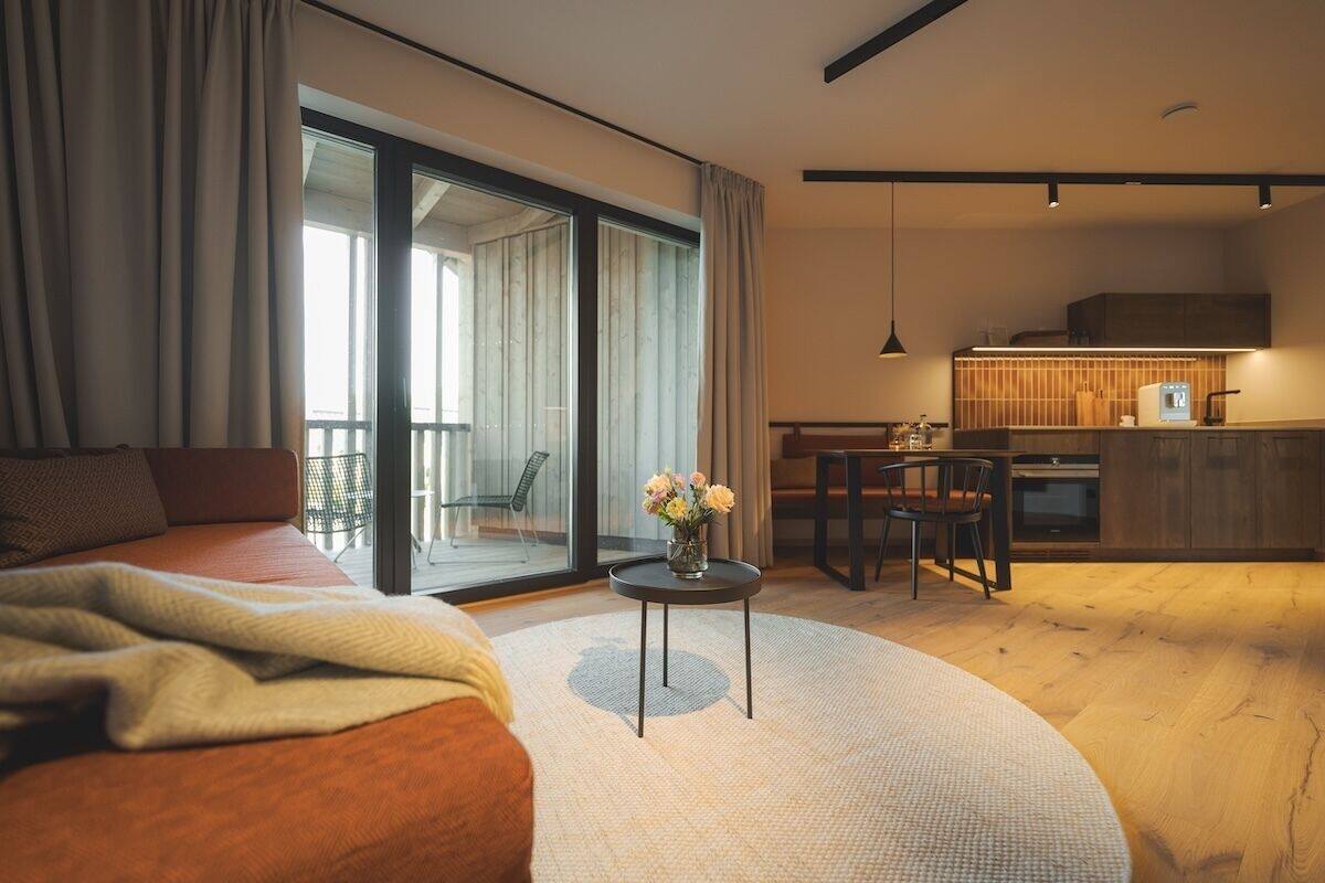 Habitación de hotel moderna con una cama grande, una mesita redonda con un florero, una cocina y un ventanal corredero que da a un balcón.