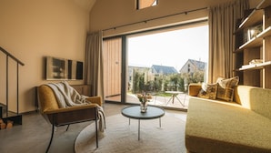 Ein modernes Wohnzimmer mit Sofa, Sessel und Couchtisch, einem an der Wand befestigten Fernseher und einem großen Fenster mit Vorhängen.