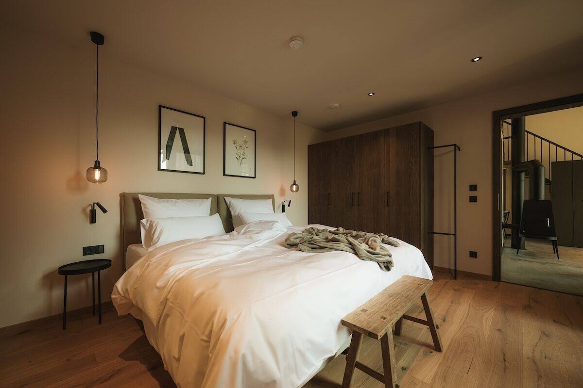 Un dormitorio moderno con una cama grande, piso de madera y un banco. Hay dos lámparas colgantes y cuadros en la pared.