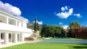 Un edificio moderno blanco con un amplio césped, una piscina y montañas al fondo.