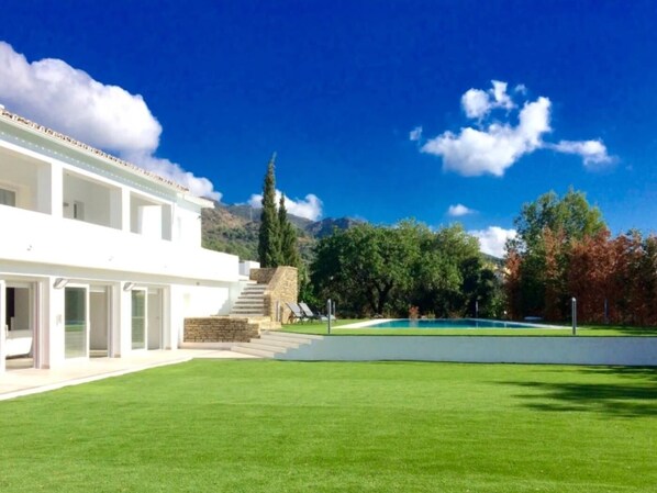 A modern white building with a large lawn, a pool, and a mountain backdrop.