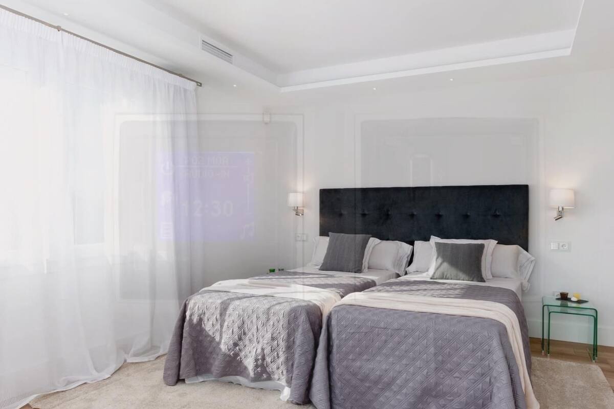 Une chambre moderne avec deux lits, une grande fenêtre avec des rideaux légers et une petite table de chevet avec une lampe.