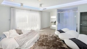 Un dormitorio moderno con cama, un ventanal con cortinas translúcidas, un sofá blanco y un box de ducha de vidrio.