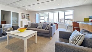 Ein modernes Wohnzimmer mit grauem Sofa, einem weißen Couchtisch und einer gelben Schale. Es gibt einen Essbereich mit Tisch und Stühlen sowie eine Küchenzeile mit Arbeitsplatte und Geräten.
