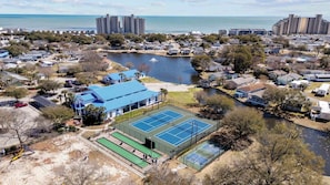 A residential area with a tennis court, a body of water, and high-rise buildings in the background. - Beautiful 4B 3.5BA in Oceanside Village - Pet friendly! 2 Golf carts included! (Surfside Beach)