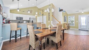 A spacious kitchen with modern appliances, a dining area with a wooden table and woven chairs, and a staircase leading to another level. - Beautiful 4B 3.5BA in Oceanside Village - Pet friendly! 2 Golf carts included! (Surfside Beach)