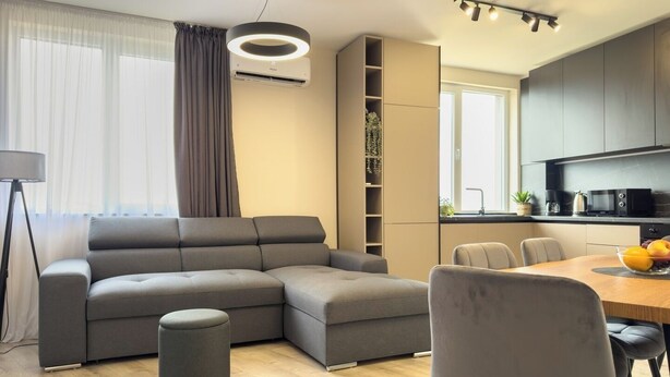 Una sala moderna con un sofá seccional gris, un taburete redondo, una mesa de comedor con sillas y una cocina con gabinetes grises y un microondas negro.