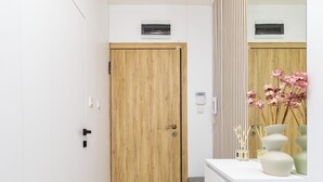 Een gang met een houten deur, een wit schoonheidsbureau met spiegel, en een vaas met roze bloemen.