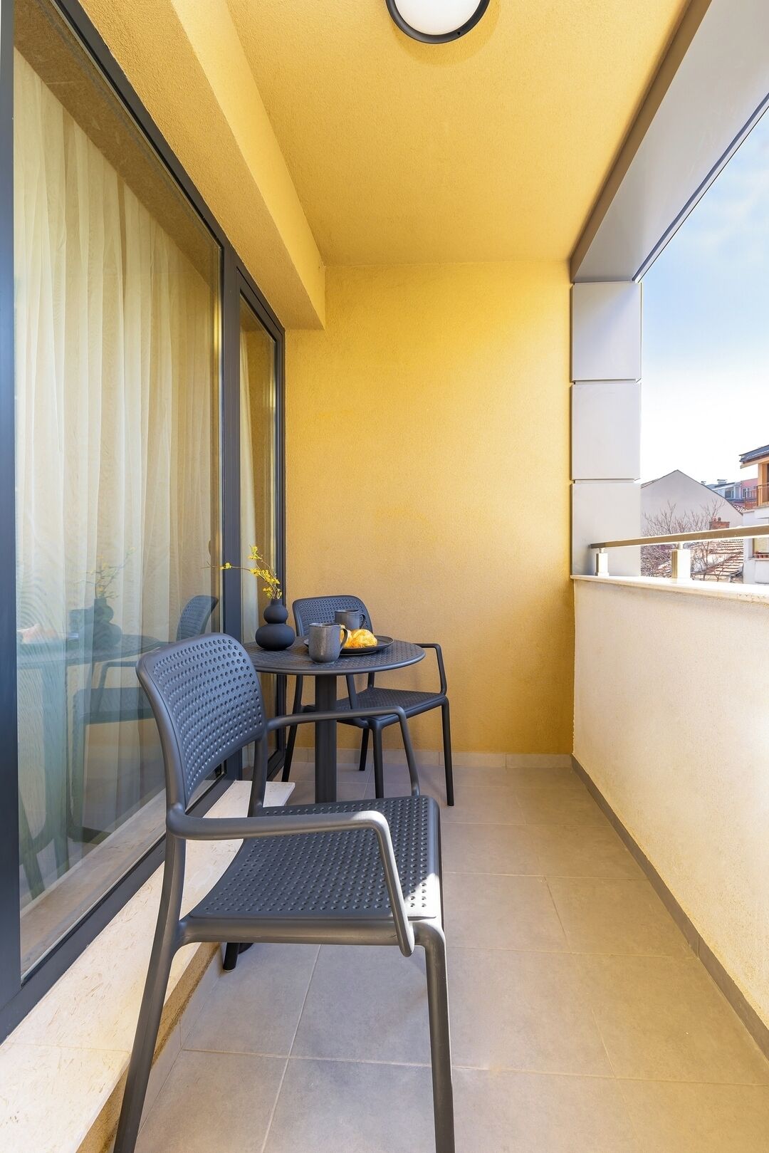 Ein Balkon mit Tisch und Stühlen, einer Blumenvase und Blick auf Gebäude im Hintergrund.