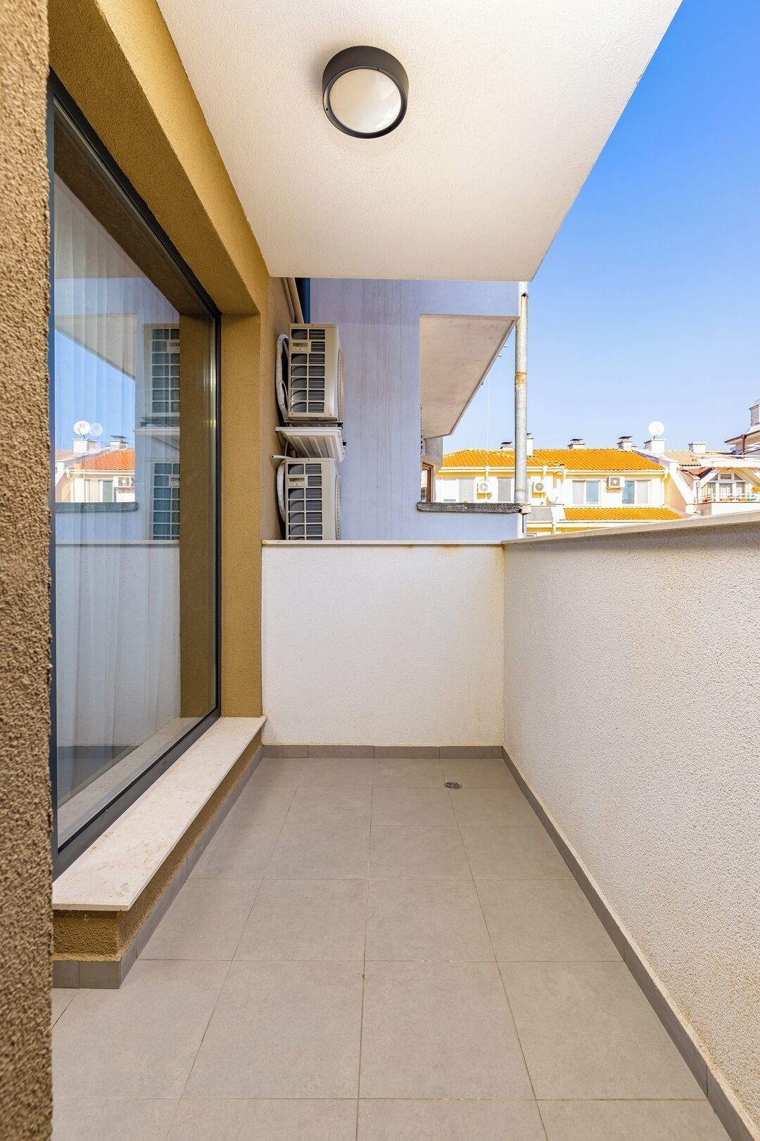 Un petit balcon avec une porte vitrée, des unités de climatisation et une vue sur les immeubles voisins.