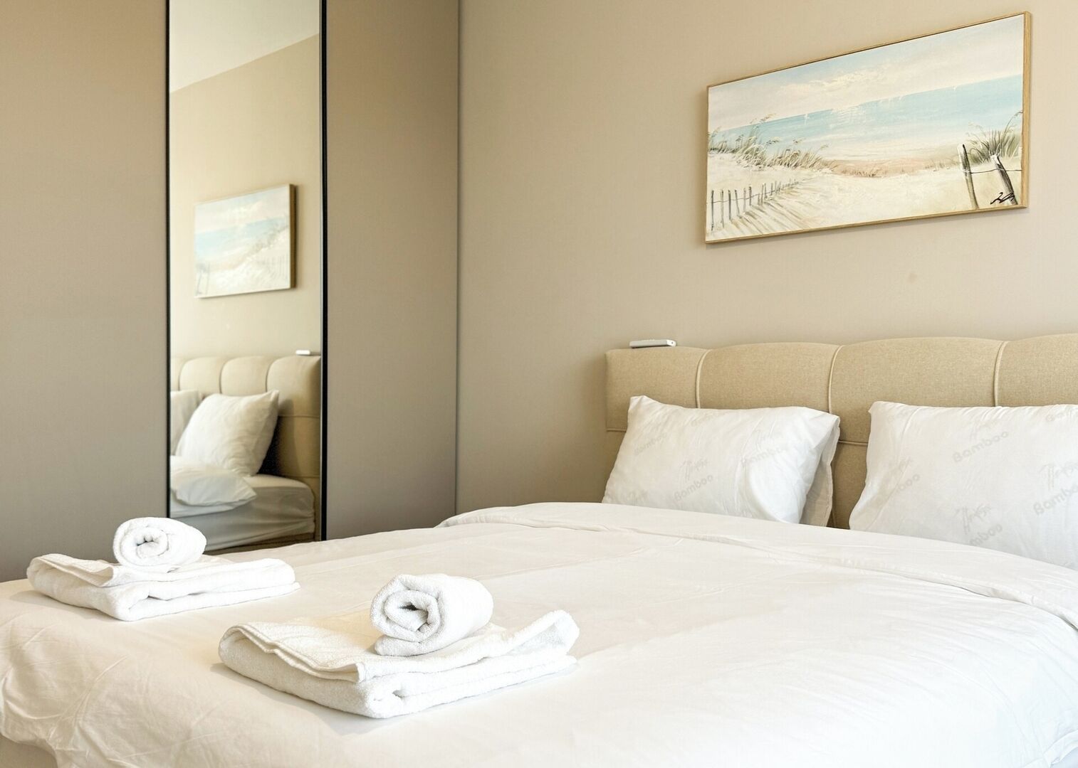 A hotel room with a large bed, two pillows, a headboard, a painting on the wall, and a mirror reflecting part of the room.