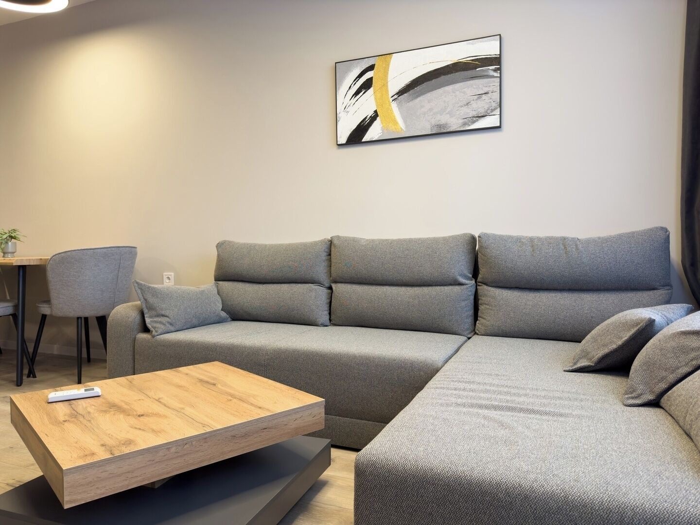 Un salon moderne avec un canapé d’angle gris, une table basse en bois et une peinture abstraite minimaliste accrochée au mur.