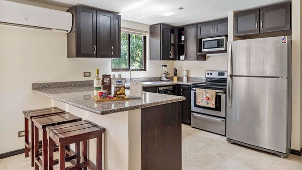 A modern kitchen with dark cabinets, a granite countertop, a stainless steel refrigerator, a microwave, an oven, and a window providing natural light. - Dual Kitchens, Pool, 3-min walk to the Beach (Playa Ocotal)