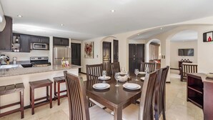 A spacious dining area with a large wooden table set for six, surrounded by dark wooden chairs. A kitchen with modern appliances is visible to the left. - Dual Kitchens, Pool, 3-min walk to the Beach (Playa Ocotal)