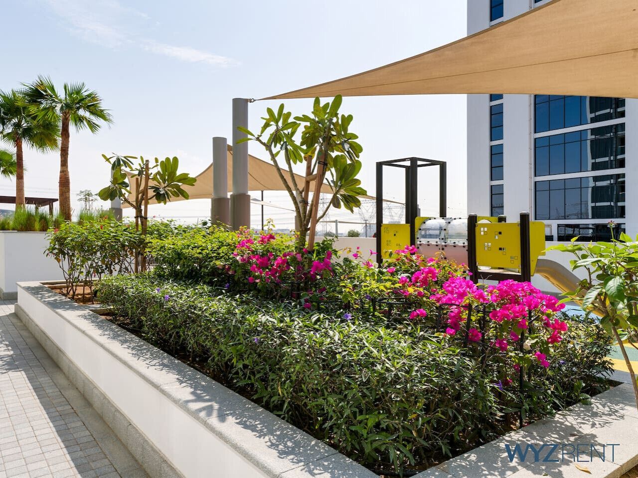 A landscaped outdoor area with a planter bed of green shrubs and pink flowers, a wooden pergola, and a modern building in the background. - Unique Studio in Azizi Riviera (Dubai)