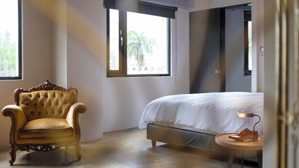 A bedroom with a large bed, a tufted armchair, a small round table with a lamp, and a window with a view of palm trees. - Tomato TTT Inn (Taipei)