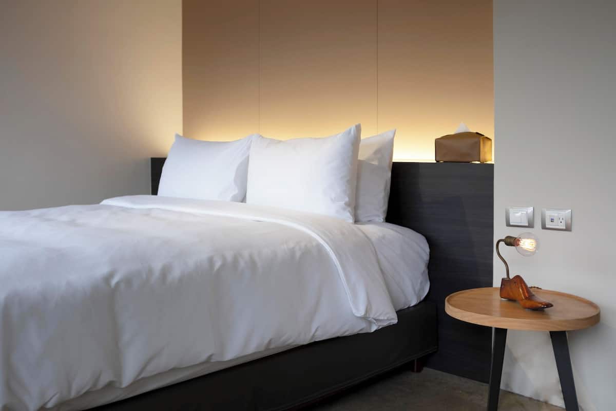 Un lit bien fait, avec du linge de lit blanc, une petite table ronde en bois avec une lampe, et une prise de courant au mur.