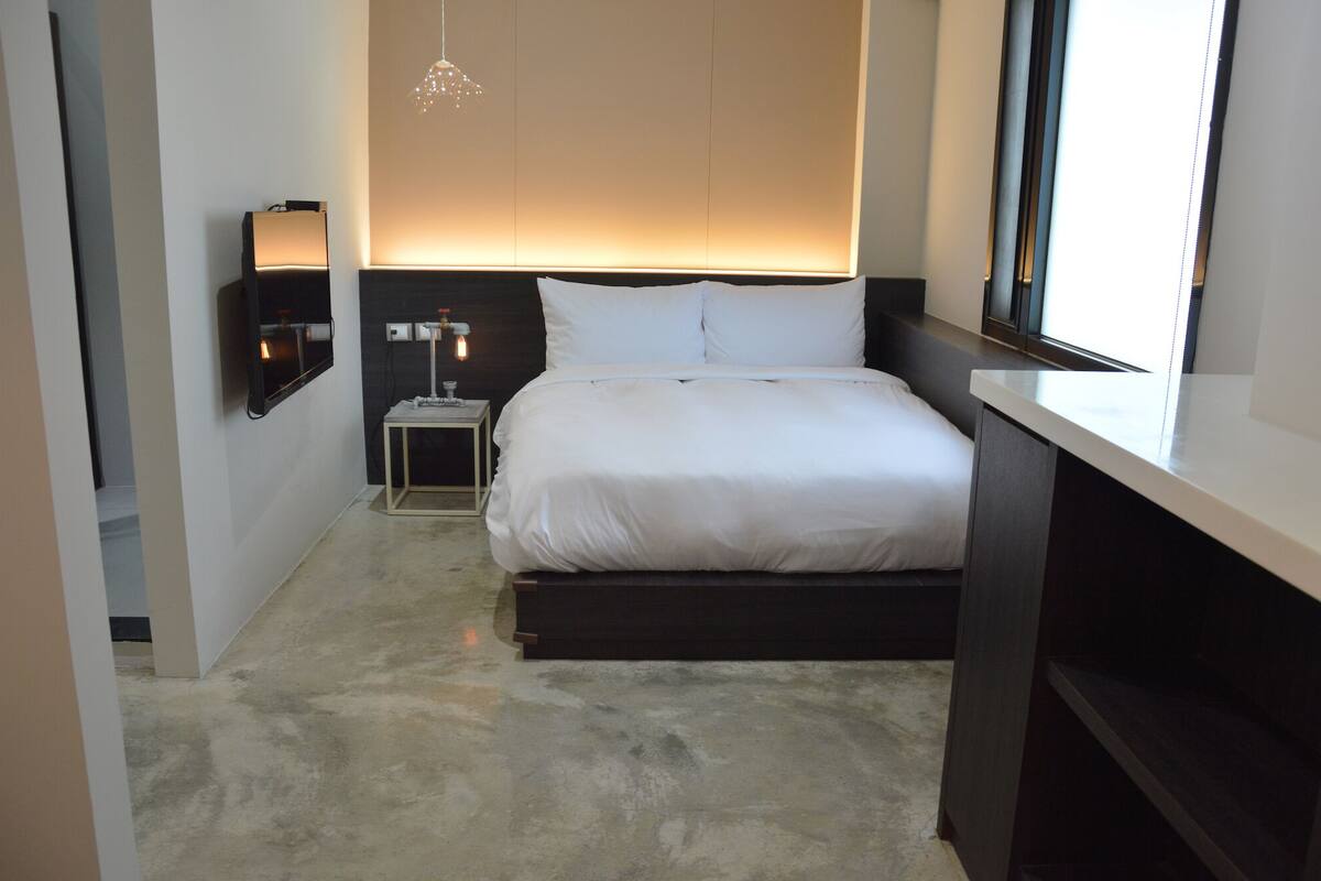 Une chambre d’hôtel moderne avec un grand lit, une table de chevet, une commode, une lampe fixée au mur et un luminaire suspendu.