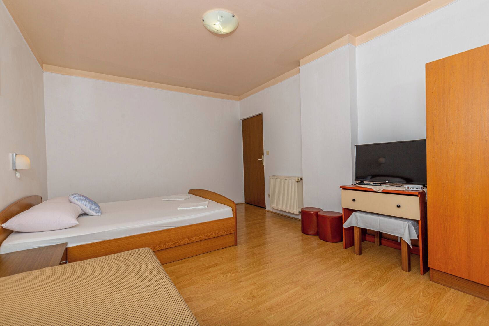 Ein kleiner, gut beleuchteter Raum mit einem Einzelbett, einem Schreibtisch mit Computer und einem Kleiderschrank.