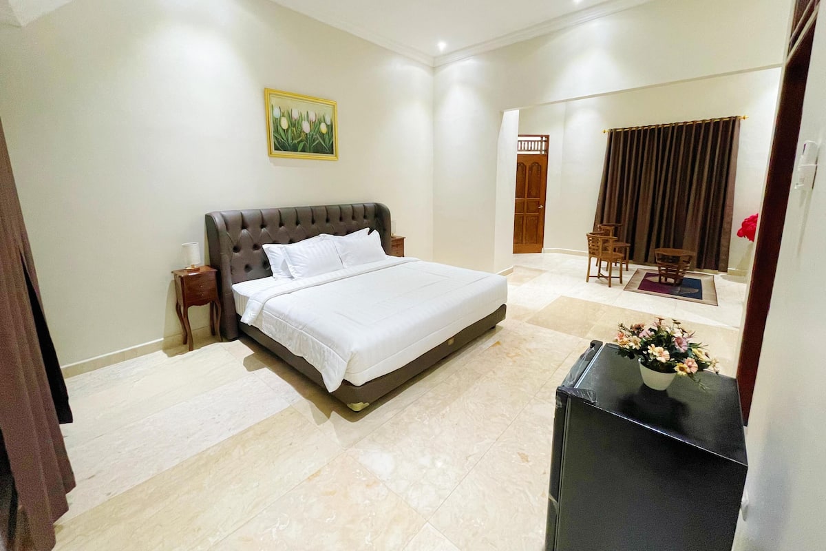 Une chambre d’hôtel avec un grand lit, une table de chevet, une commode, un bureau avec une chaise, une plante en pot et une porte donnant sur une autre pièce.