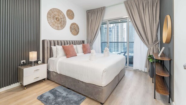Een moderne slaapkamer met een groot bed, een nachtkastje met een lamp, en uitzicht op hoge gebouwen door het raam.