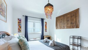 Ein ordentlich bezogenes Bett mit Kissen, ein Nachttisch mit einem Schiffsmodell, eine hängende Chandelier, ein Fenster mit Vorhängen und eine Wandteppich.