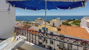Ein Balkon mit einem blauen Sonnenschirm, einem Tisch mit Gläsern und einer Flasche sowie Blick auf das Meer und Gebäude.