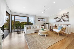 A spacious living area with a white sofa, a round wicker ottoman, a wicker chair, a ceiling fan, and a large window offering a view of greenery.