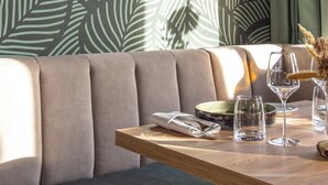 A restaurant interior with a leaf-patterned wall, a wooden table with glasses and a vase, and a sofa in the foreground. - Feinheit Hotel & Restaurant (Halsenbach)