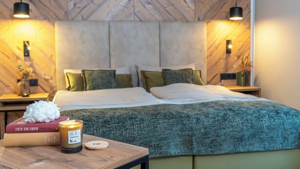 A modern bedroom with a herringbone wood ceiling, a bed with green and white bedding, a bedside table with a candle, books, and a decorative item. - Feinheit Hotel & Restaurant (Halsenbach)
